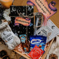 A gift box containing a 'ROMANTASY - SPICY FANYASY' paperback book, cozy reading socks, annotation tabs, a highlighter pen, bookmarks, handmade stickers, a Pukka tea bag, and options hot chocolate and Milky Way Magic Stars chocolates.