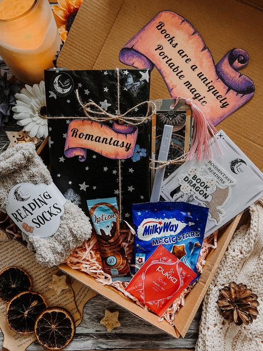 A gift box containing a 'ROMANTASY - SPICY FANYASY' paperback book, cozy reading socks, annotation tabs, a highlighter pen, bookmarks, handmade stickers, a Pukka tea bag, and options hot chocolate and Milky Way Magic Stars chocolates.
